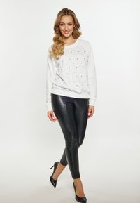 Witte sweatshirt met zilveren versieringen, gecombineerd met strakke zwarte faux leren broeken en zwarte hoge hakken, wat een strakke, moderne look creëert.