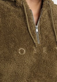 Olivgrön fleece-hoodie med dragkedja halvvägs, texturerad tyg och broderad text framtill. Har en huva.
