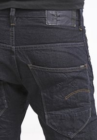 Primer plano de un bolsillo trasero de unos jeans de mezclilla oscura con costuras marrones y una etiqueta de cuero negro de la marca sobre la cinturilla.