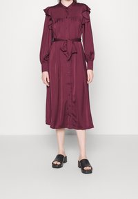 Burgundské šaty s dlouhým rukávem, zdobené volánky, s knoflíkovým zapínáním v přední části a opaskem uvázaným v pase; hladký materiál. V kombinaci s černými robustními sandály.