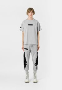 Camiseta gris y pantalones de chándal a juego con diseños negros similares a plumas. La camiseta presenta etiquetas; los pantalones de chándal tienen bolsillos laterales y puños.