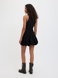 Črn rebrast brezrokavnik v kombinaciji z obsežno črno krilo, dopolnjeno z visokimi črnimi čevlji do kolen, ki imajo zaponke. Gladke teksture skozi celotno obleko.