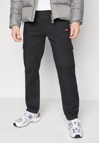 Svarta cargo byxor med flera fickor, tillverkade av hållbart tyg. Bärs med en grå puffjacka och vita sneakers med röda detaljer.