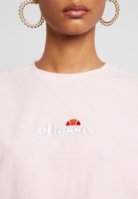 Gai rozā kokvilnas T-krekls ar apaļu kakla izgriezumu, kuram ir izšūts "ellesse" logo un pusapļa dizains sarkanā un oranžā krāsā virs teksta.