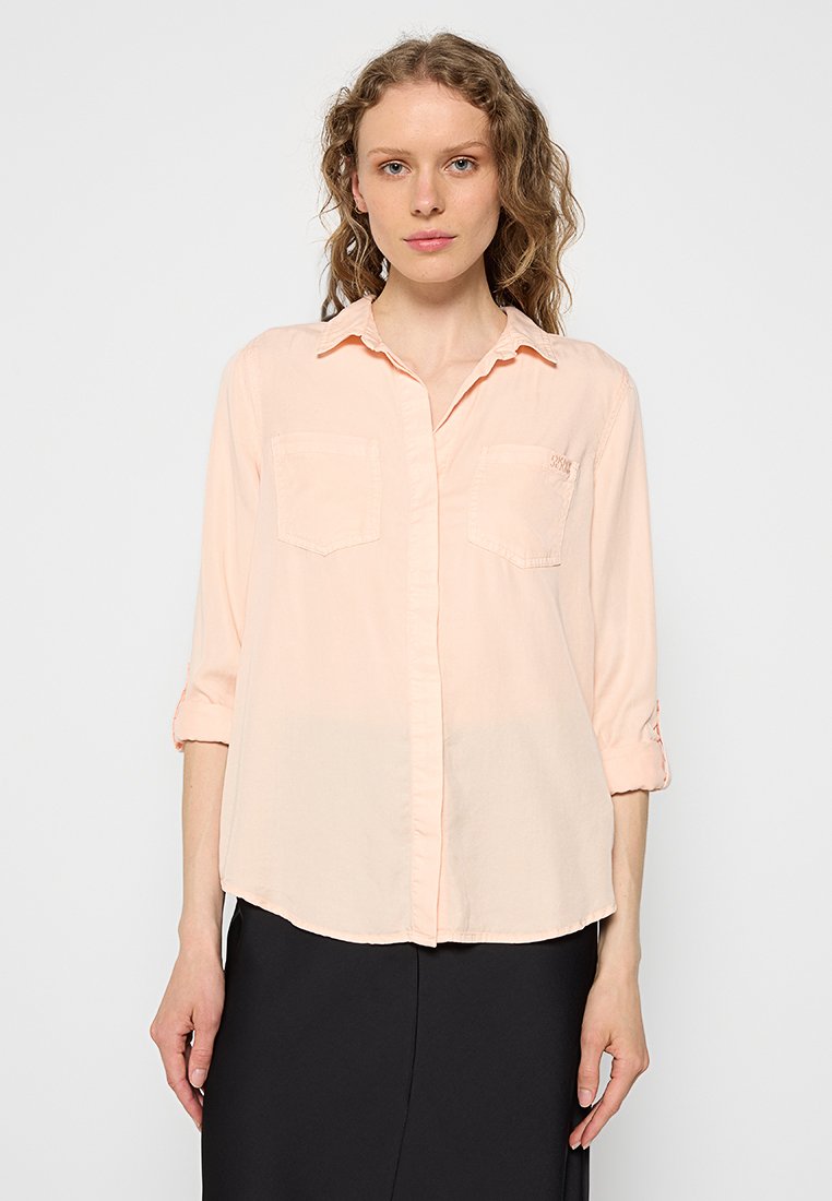 Camisa de botones rosa claro con una textura suave, que cuenta con dos bolsillos en el pecho y mangas enrolladas, fabricada con material ligero.