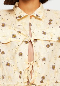 Chemise boutonnée à motif floral en tissu jaune clair, avec des fleurs violettes et des motifs à points, dotée d'une fermeture à nouer et d'un détail d'encolure dégagée.