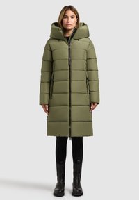 Olivgrüner Puffer-Mantel mit Kapuze, ausgestattet mit einem Reißverschluss, seitlichen Taschen und einem gesteppten Muster. Wird über schwarzen Leggings und hohen Stiefeln getragen.