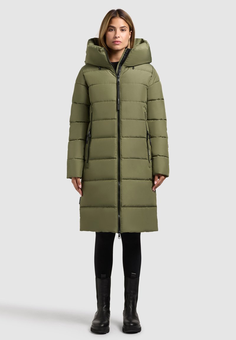 Olivgrüner Puffer-Mantel mit Kapuze, ausgestattet mit einem Reißverschluss, seitlichen Taschen und einem gesteppten Muster. Wird über schwarzen Leggings und hohen Stiefeln getragen.
