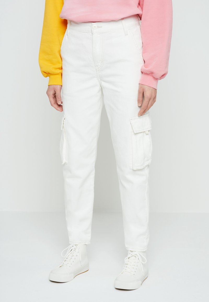 Personne portant un pantalon cargo blanc, des baskets hautes blanches et un sweat-shirt aux manches jaunes et roses, debout sur un fond blanc.