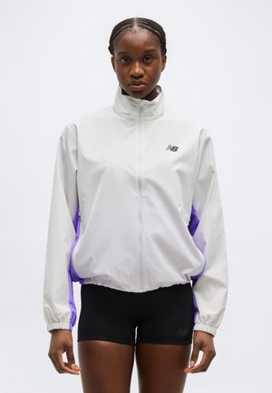 Mujer con cabello trenzado, vestida con una chaqueta blanca y morada con cremallera y pantalones cortos deportivos negros, de pie frente a un fondo blanco liso.