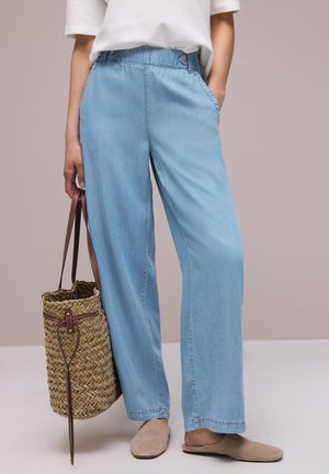 Person iført lyseblå bukser med vide ben, hvid top med korte ærmer, beige slip-on sko og med en flettet kurvetaske med brune stropper.