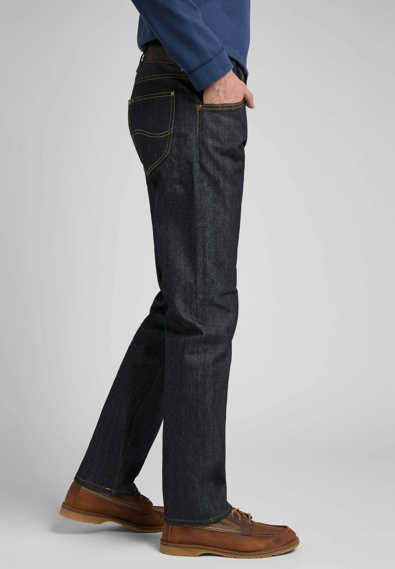 Jeans de mezclilla oscura con un corte recto, acentos de costuras amarillas y bolsillos traseros. Combinados con zapatos marrones de cordones y una camisa azul de manga larga.