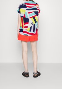 Top corto a maniche corte colorato con motivi geometrici astratti e testo, abbinato a pantaloni corti arancioni vivaci e sandali neri con cinturini logo.