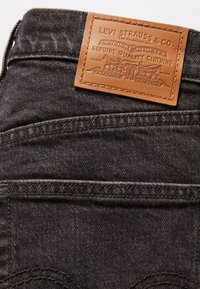 Jean en denim noir avec une étiquette en cuir beige cousue Levi Strauss & Co. sur la ceinture au-dessus de la poche arrière.