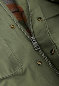 Olivgrüne Jacke mit einem silbernen Reißverschluss, goldenen Druckknöpfen und einer strukturierten Stoffauskleidung mit einem Karomuster darunter.