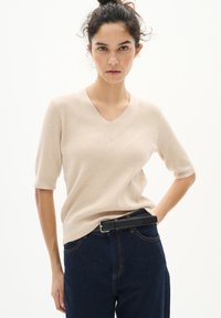 Beige ribbad kortärmad V-ringad tröja, kombinerad med högmidjade mörka denimjeans och ett svart läderbälte. Slät textur genom hela plagget.