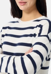 Sweterek w paski w kolorach kremowym i granatowym, o miękkiej fakturze, okrągłym dekolcie i opadających ramionach. Pokazuje górną część ciała z rękoma skrzyżowanymi.