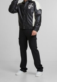 Veste en cuir noire avec accents blancs, boutons-pression et détails de logo. Associée à un pantalon cargo noir et des baskets blanches.