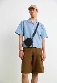 Lyseblå kortermet skjorte med logo, brune cargo-shorts og en svart rund crossover-veske. Tilbehør inkluderer et sølvarmbånd og caps.