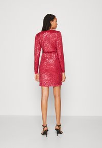 Robe courte en sequins rouges avec des manches longues, coupe ajustée, taille ceinturée et décolleté rond. Portée avec des sandales à talons hauts noires.