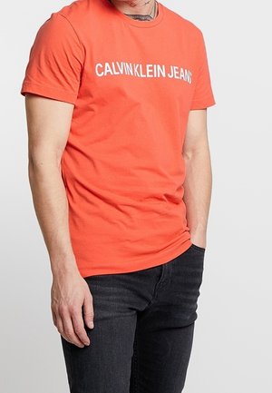 Mężczyzna w jaskrawo pomarańskiej koszulce Calvin Klein Jeans i czarnych dżinsach, z jedną ręką w kieszeni, na jednolitym tle.