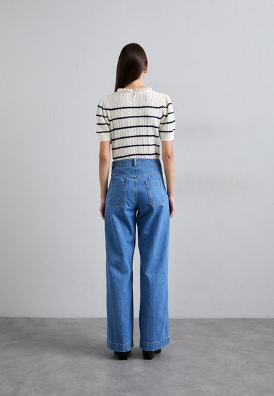 Pleteni sveten s krem in mornarsko modrimi črtami s kratkimi napihnjenimi rokavi, v kombinaciji z visokim pasom in širokimi modrimi jeans hlačami, pogled odzadaj.