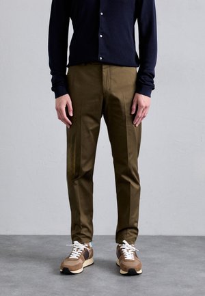 Homme portant un pantalon vert olive, des baskets marron et beige, et une chemise noire à manches longues boutonnée, debout sur un sol gris.