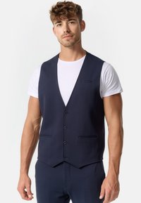 Chaleco de color azul marino con una textura suave, que presenta un escote en V profundo, cinco botones y dos bolsillos frontales. Se lleva sobre una camiseta blanca.