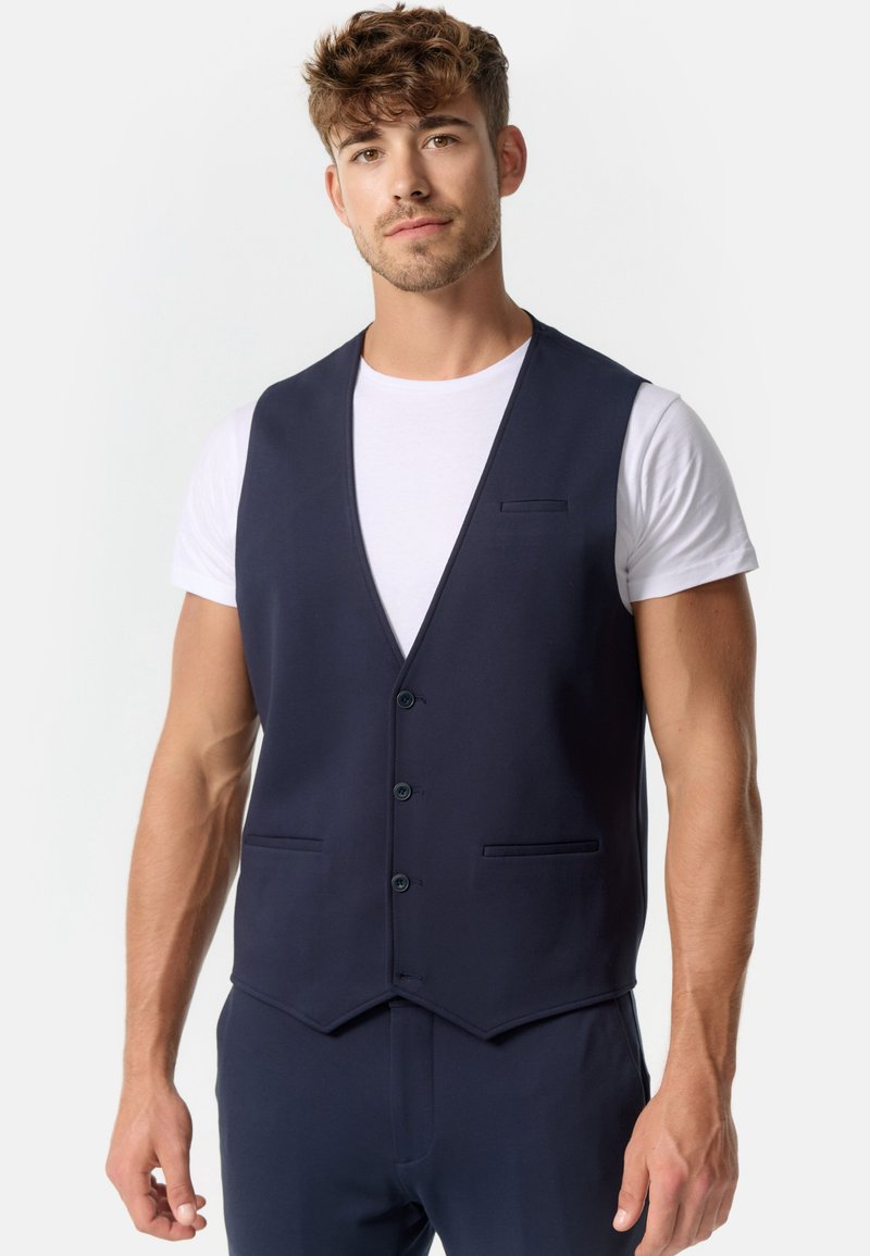 Chaleco de color azul marino con una textura suave, que presenta un escote en V profundo, cinco botones y dos bolsillos frontales. Se lleva sobre una camiseta blanca.
