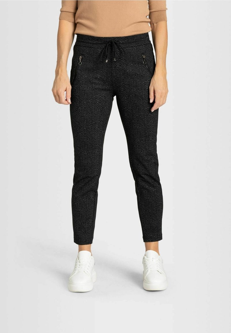 Kvinna i svarta strukturerade byxor med dragkedjefickor, en beige topp och vita sneakers, stående mot en enfärgad bakgrund.