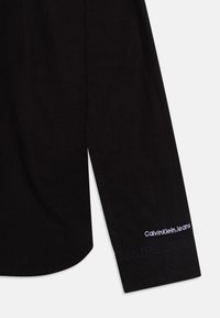 Svart skjorta med långärmar, med en böjd fåll och broderad "Calvin Klein Jeans"-logotyp i vitt på ärmslutet. Slät tygtextur.
