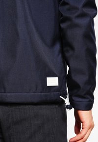 Giacca blu navy in tessuto liscio con orlo elastico, etichetta bianca in basso e coulisse laterale per la regolazione.