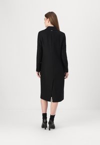 Cappotto in lana nera con silhouette dritta, design doppiopetto, colletto a scollo a V e spacco sul retro. Abbinato a scarpe con tacco alto.