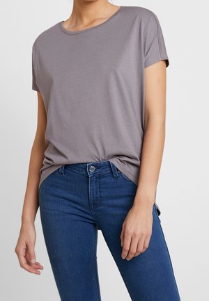 Kvinde iført en grå kortærmet T-shirt stukket ned i blå jeans, stående med den ene hånd afslappet ned langs siden.