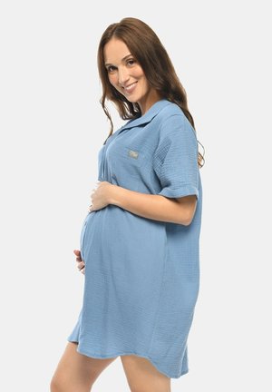 Schwangere Frau lächelt, trägt ein lockeres blaues Kleid, hält ihren Bauch mit beiden Händen und steht vor einem einfachen, hellen Hintergrund.