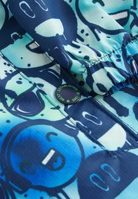 Färggamt badklädestyg med blå och teal tecknade mönster, med en rynkad midja och en svart knapp märkt "Etablerad 1982."