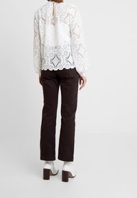 Femme de dos portant un chemisier en dentelle blanche à manches longues, un pantalon droit marron foncé et des bottines blanches à talons, sur un fond blanc.