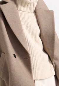 Beige wollen jas met een op maat gemaakte kraag over een crèmekleurige geribde trui. De jas heeft een zwarte knop en een zachte textuur.
