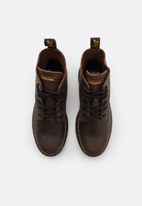 Paire de bottes Dr. Martens en cuir brun foncé avec lacets noirs et languettes de tirage jaunes marquées, vue de dessus sur un fond gris.