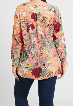Femme portant un chemisier pêche à manches longues avec un motif floral coloré, vue de dos, associée à un jean bleu foncé.