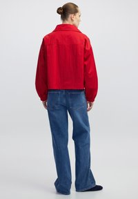 Vrouw met een knot in haar haar, gekleed in een losse rode jas en wijde blauwe jeans, die met de rug naar de camera staat tegen een effen lichte achtergrond.