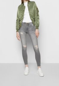 Olivgrön bomberjacka med en glänsande yta, åtsittande vit top, slitna grå skinny jeans och vita sneakers.