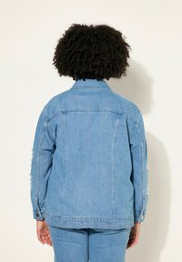 Giacca in denim azzurro chiaro con un design scucito, polsini con bottoni e colletto classico. Cuciture visibili e tasche sul retro.