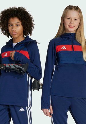 Chaquetas deportivas en color azul marino con acentos rojos, que incluyen cremalleras y paneles laterales. El modelo a la izquierda sostiene zapatos de fútbol; ambos llevan pantalones a juego.