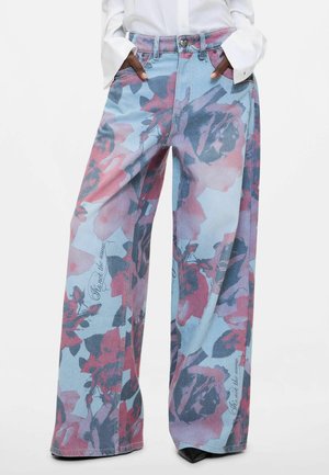 Wijde blauwe spijkerbroek met grote roze en donkere bloemmotieven en cursieve tekst, gedragen met een wit shirt met lange mouwen en zwarte puntige schoenen.