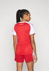 Maillot de sport rouge avec des manches blanches, présentant un motif texturé. Associé à un short rouge. Vue arrière, mettant en avant le logo au dos.