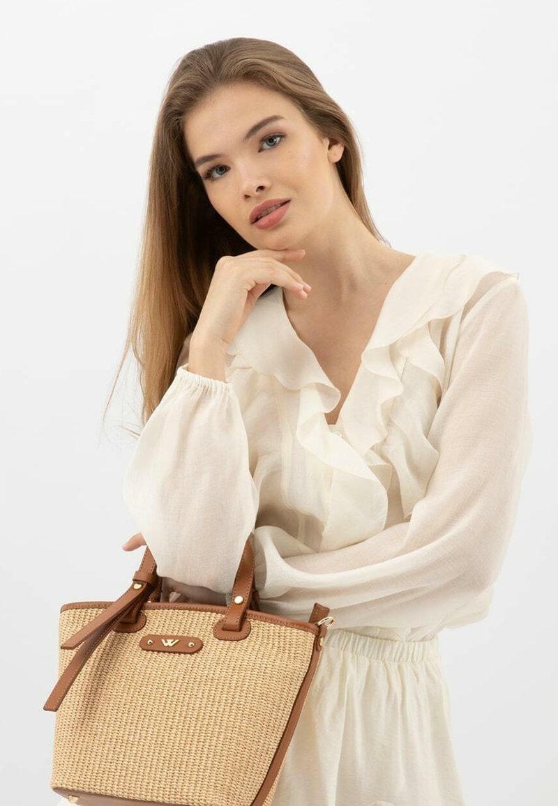 Słomiana torebka z brązowymi skórzanymi akcentami, wyraźnym kształtem, krótkimi uchwytami i złotymi detalami. Modelka nosi kremową, falbanistą bluzkę.