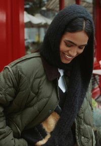 Gepolsterte olivgrüne Bomberjacke mit einem braunen Cordkragen, über einem gemusterten schwarz-beigen Pullover getragen, ergänzt durch eine schwarze Kapuze.
