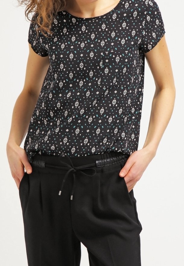 Femme portant un haut à manches courtes noir à motifs, rentré dans un pantalon noir à cordon, les mains dans les poches.