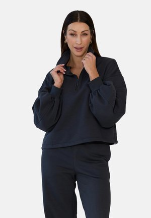 Kobieta o długich brązowych włosach, nosząca złote kolczyki oraz luźną czarną bluzę na zamek i pasujące spodnie, trzymająca kołnierz obiema rękami.